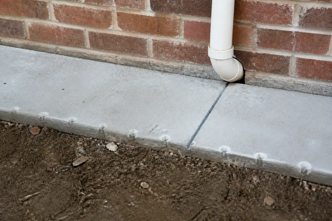 The base of a brick wall where a concrete slab meets grade, showing an expansion joint and a plumbing penetration